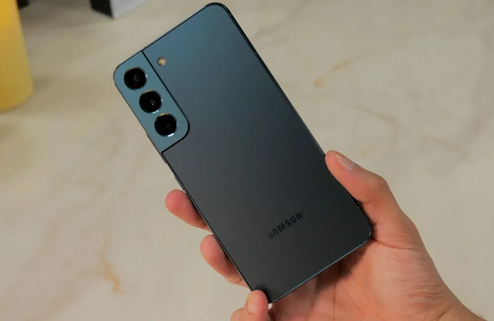 Рука держит тёмно-зелёный или чёрный смартфон Samsung Galaxy S22, показывая его заднюю панель с тройной камерой. На нижней части корпуса виден логотип «SAMSUNG»