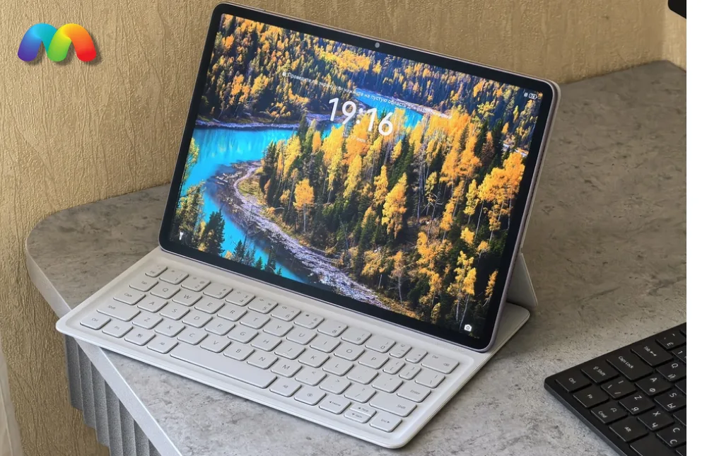 Планшет Huawei MatePad 11.5 2025 установлен на белую клавиатуру и стоит на столе. На экране отображается яркий пейзаж с рекой и лесом.