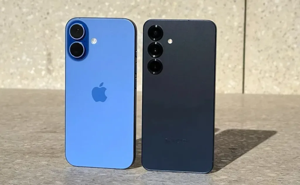 Черный Samsung Galaxy S25 и синий iPhone 16 стоят рядом на столе, демонстрируя свои задние панели с камерами.