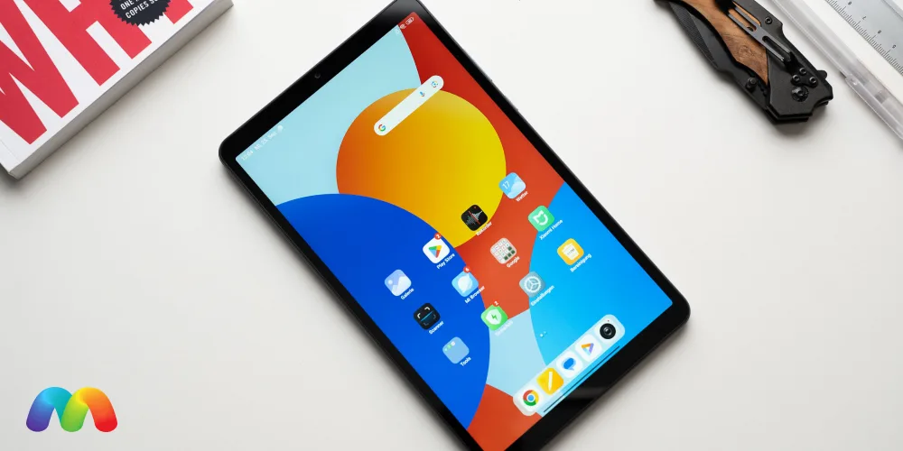 Планшет Xiaomi Redmi Pad SE 8.7, лежащий на белом столе, с видимым рабочим столом, иконками приложений и яркими обоями. На заднем плане видны книга и пенал.
