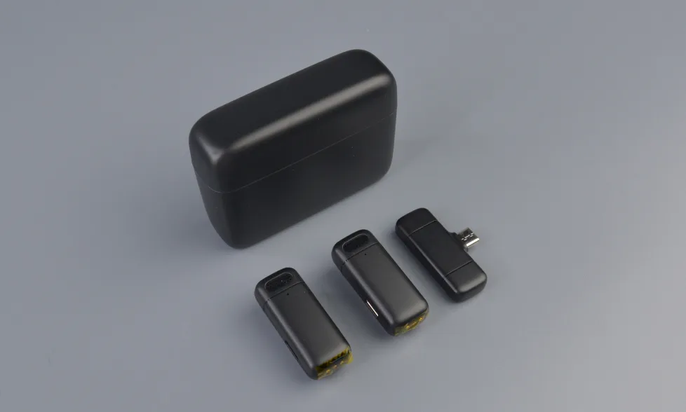Черный зарядный чехол и три черных миниатюрных устройства беспроводного микрофона (два передатчика и один приемник) на сером фоне.